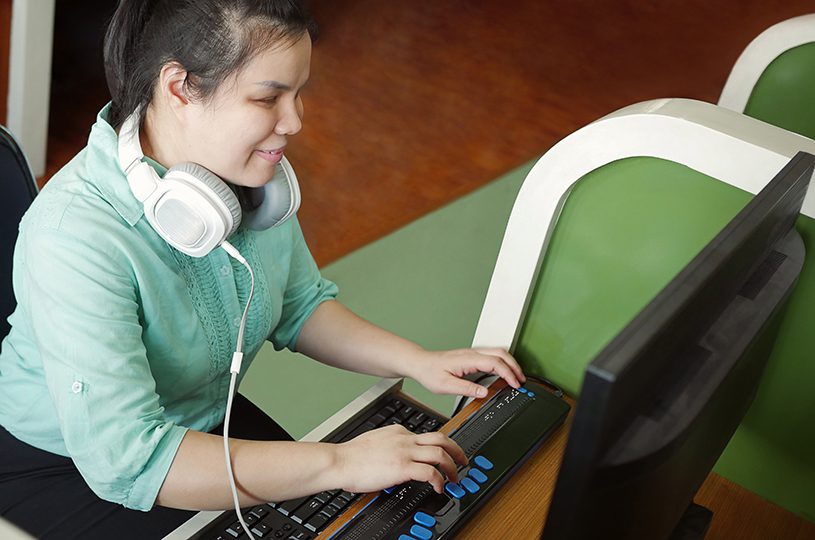 Asian young blind woman with headphone using computer with refreshable braille display or braille terminal a technology device for persons with visual disabilities.